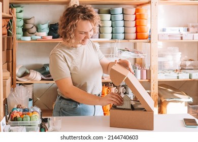 Smiling Blond Curly Woman Packing And Wrapping Shipment With Postcards, Silicone Baby Dishes In Cardboard Delivery Box. Colorful Skein Of Thread In Container On Desk. Workshop Store, Small Business