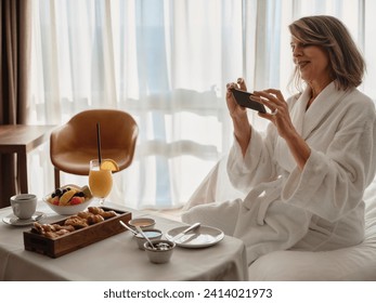 Smiling blond elderly woman photographing breakfast through smart phone while sitting on bed in hotel room - Powered by Shutterstock