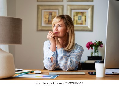 Smiling blond haired businesswoman deep in thought while sitting at desk and looking out the window. Home office. 