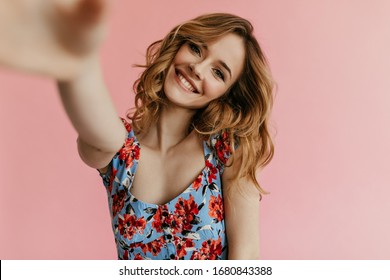 Smiling blond haired woman in blue floral top makes photo on isolated background. Cool girl with curly hair takes selfie..