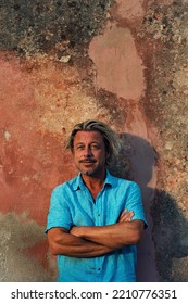 Smiling Blond Man With Crossed Arms In Blue Shirt Stands Against Weathered Pink Plaster Wall In Late Afternoon Sunlight.