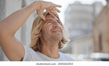 A smiling blond man with long hair shades his eyes while looking upwards on a sunny urban street. - Powered by Shutterstock