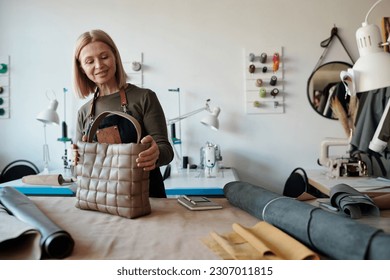 Smiling blond mature craftswoman with stylish handbag standing by workplace and preapring new leatherwear item for packing - Powered by Shutterstock