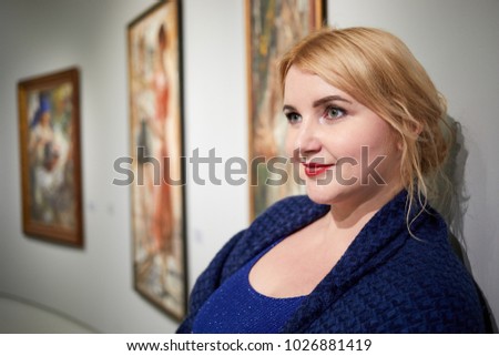 Smiling blond woman in blue dress stands against paintings on wall.