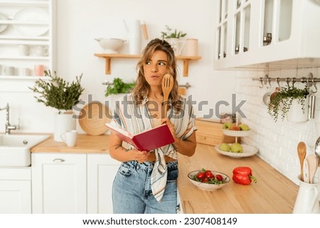 Smiling blond woman looking at recipe in cookery book and holding apple in light kitchen at home. Dieting healthy lifestyle concept.