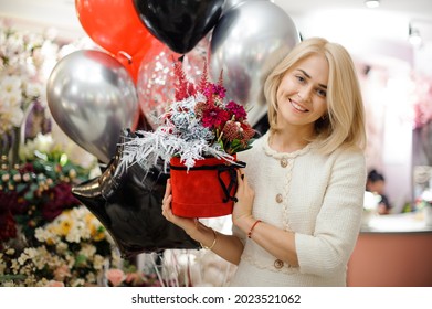 Smiling Blond Woman With Round Box Of Flower Arrangement In Her Hands. Floral Shop Concept. Flowers Delivery. Red And Silver Colors.