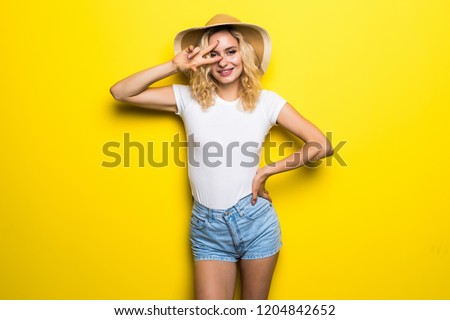 Smiling blond young woman with victory sign in straw hat, jeans shorts, white shirt on yellow background.