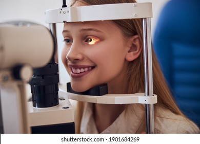 Smiling Blonde Caucasian Female Child In Blue Medicine Chair And Looking To Slit Lamp Machine During Medical Check Up In Eyes Clime