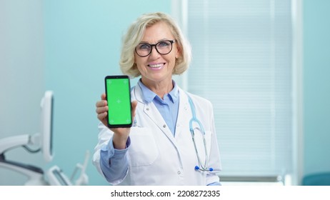 Smiling Blonde Doctor Shows Results Of Analysis Holding Phone In Hand. Woman Makes Appointment To Ultrasound Diagnostics Via Chromakey Green Screen