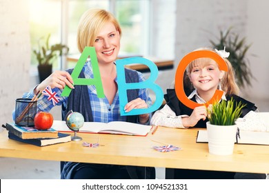 Smiling blonde English teacher plays letters and words with kid school girl, table with books in light classroom with flowerpots