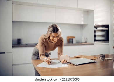 A smiling blonde filling up bills while standing in kitchen at her cozy home. - Shutterstock ID 2121075095