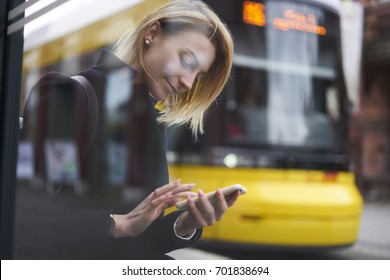 Smiling blonde hipster girl waiting for public transport on bus stop chatting in social networks via smartphone,casually dressed female tourist using application for searching traffic schedule - Powered by Shutterstock