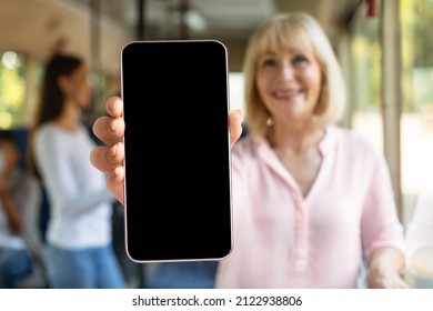 Smiling Blonde Mature Lady Taking Bus Holding Cell Phone With Black Blank Screen In Hand, Showing Device Close To Camera. Gadget With Empty Free Space For Mock Up. Online Ticket, Gps Navigation