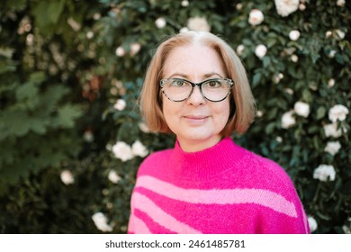 Smiling blonde mature woman 50-60 year old with gray hair wearing vision glasses and pink knitted sweater over blooming rose flowers outdoor. Looking at camera.  - Powered by Shutterstock