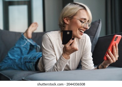 Smiling Blonde Mid Aged Woman Shopping Online With Credit Card And Mobile Phone On A Couch At Home