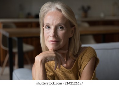 Smiling blonde middle aged 60s lady home head shot portrait. Skinny retired woman, pensioner, OAP sitting on couch, touching chin, looking at camera, posing for profile picture