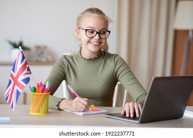 Smiling Blonde School Girl With Flag Of Great Britain Using Laptop, Having Educational Online Course, Studying English On Internet, Home Interior. English Classes Online Concept