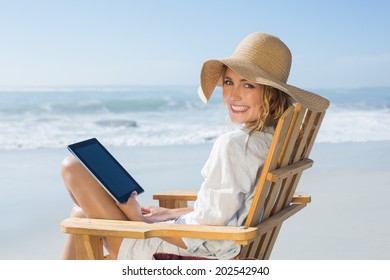 Smiling blonde sitting on wooden deck chair by the sea using tablet on a sunny day