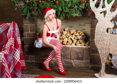 Smiling Blonde Wearing Red Christmas Robe, Shoes, Hat And Striped Knee Socks Sits Near Hearth In Drawing Room