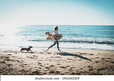 Smiling blonde woman running alongside the sea side with her pet dog on a sunny day. Cheerful Caucasian female in blue jeans with plaid on her shoulders playing with her doggy at the sandy beachside. - Powered by Shutterstock