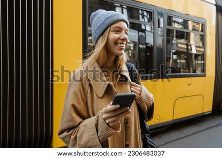 Smiling blonde woman wearing warm hat using mobile phone while standing at bus station
