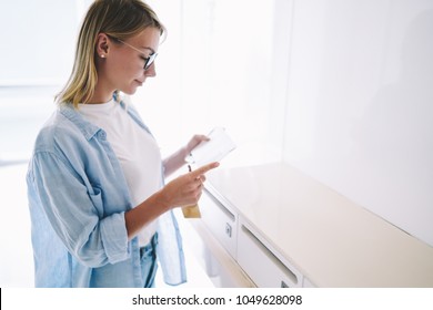 Smiling Blonde Young Woman Dressed In Casual Clothes Reading Letter With Good News.Cheerful Hipster Girl In Eyewear Getting Envelope With Invitation By Mail Standing In Corridor Near Postbox