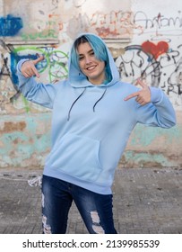 Smiling Blue Haired Teenager Pointing In Center Of Blue Hoodie. Hooded Teen Girl Stays Outdoors Against Graffiti Wall. Clothing Mockup 