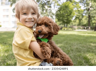 smiling boy of 6-7 years old sits on the grass with a fluffy brown poodle puppy. Face portrait of a boy with a dog in the summer for a walk. Furry friend, dream of having a dog - Powered by Shutterstock