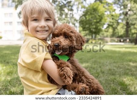 smiling boy of 6-7 years old sits on the grass with a fluffy brown poodle puppy. Face portrait of a boy with a dog in the summer for a walk. Furry friend, dream of having a dog
