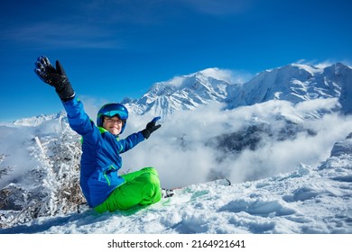 Smiling boy with alpine ski sit in snow lifting hands, wear helmet and sport glasses on top of the mountain over peaks covered by clouds on background