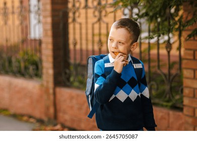 Smiling boy with backpack eats sandwich outdoors before school, morning routine, healthy snack, back to school season