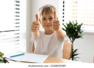 A smiling boy does his homework at the table and gives a thumbs up. Distance learning online education. Elementary school. - Powered by Shutterstock