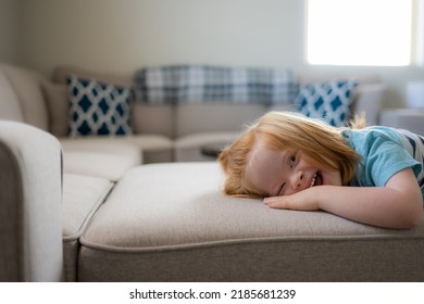Smiling boy with Down syndrome laying his head on light colored sofa backlit by window