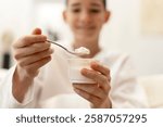 Smiling boy eating yogurt from a plastic cup with a spoon, enjoying a healthy snack or breakfast at home, promoting healthy eating habits for children