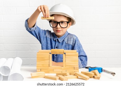 Smiling Boy Engineer In A Construction Helmet And Glasses Builds A House From Wooden Blocks With Plans And A Measure Tape. Child Builder Plays With Cubes At Home. Mortgage, Investment, Real Estate.