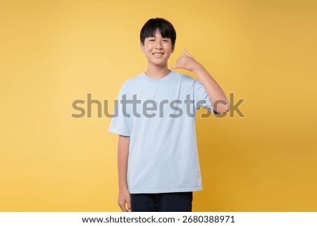 Smiling boy gesturing "call me" in front of a yellow backdrop wearing a light blue t-shirt