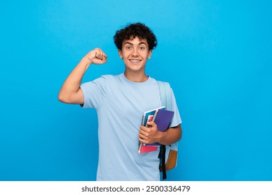 Smiling boy guy high school male pupil student teenager with backpack holding books copybooks winning gesture celebrating having success standing isolated on blue background copy space