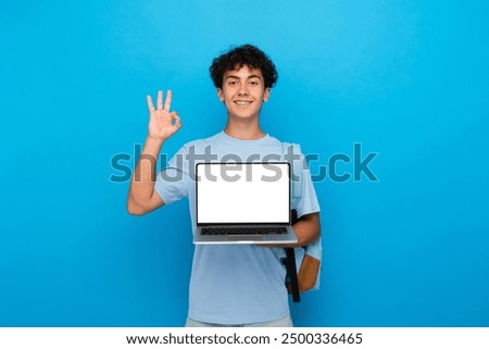 Smiling boy guy high school male pupil student teenager with bag holding using laptop computer for e-learning showing okay gesture sign demonstrate mock up screen isolated over blue background