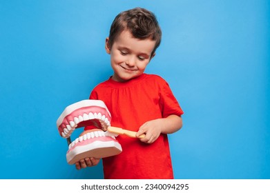 Un niño sonriente con dientes sanos sostiene una mandíbula grande y un cepillo de dientes en sus manos sobre un fondo azul aislado. Higiene oral. Odontología pediátrica. Reglas para cepillar dientes.