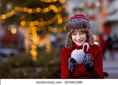 Smiling Boy Holding Cup With Hot Chocolate, Marshmallows And Candy Canes Enjoying Outdoor Holiday Time With Decorated With Lights Christmas Tree In The Background, Copy Space On Left, Family 