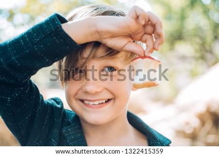 Smiling boy holds bitten apple with one hand. The boy advertises children's clothes for the autumn. Cute boy on autumn holiday in village. Happy childhood. Joyful guy with hat on his head in the hay