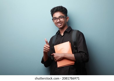 Smiling boy of Indian ethnicity holding note books in his hands and shows thumbs up gesture