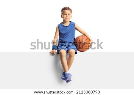 Smiling boy in a jersey holding a basketball and sitting on a blank panel isolated on white background