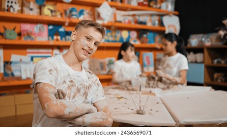 Smiling boy looking at camera and crossing arm with confident at workshop while diverse student having pottery class together. Happy caucasian student smile while pose with arm folded. Edification. - Powered by Shutterstock