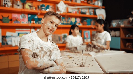 Smiling boy looking at camera and crossing arm with confident at workshop while diverse student having pottery class together. Happy caucasian student smile while pose with arm folded. Edification. - Powered by Shutterstock