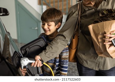 Smiling Boy Looks How Mom Charges Electric Car At Home With Charger Plug Handle