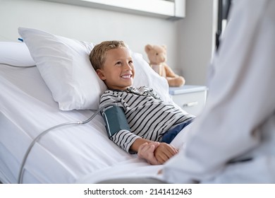 Smiling Boy Lying On Hospital Bed Getting Blood Pressure Checked. Good Child Patient Looking At Doctor While Holding Hand During Check Up At Clinic. Strong Happy Kid Recovering From Sickness.