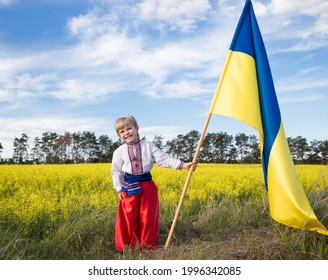 Smiling boy in national Ukrainian clothes with yellow - blue large flag in hand against background of blooming rapeseed field. pride, symbol of country, nationality of Ukraine. Patriotic education.