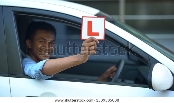 Smiling boy showing l-plate at car window,\
driving courses, right-hand\
traffic