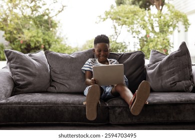 Smiling boy using tablet on cozy sofa in sunny outdoor setting. Technology, outdoors, relaxation, happiness, childhood, leisure
